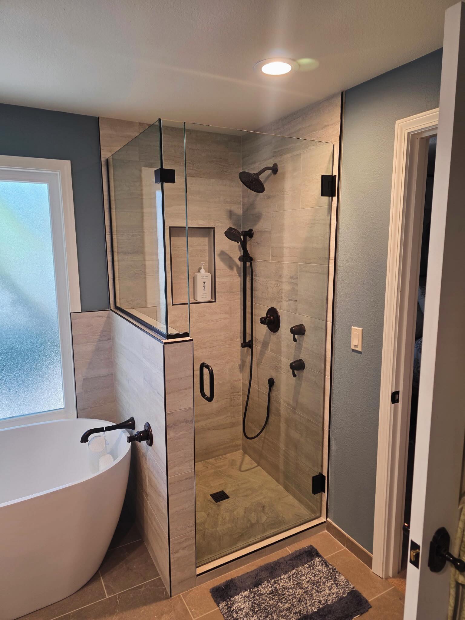 Finished bathroom with glass shower, gray tile, dark fixtures and a freestanding tub.